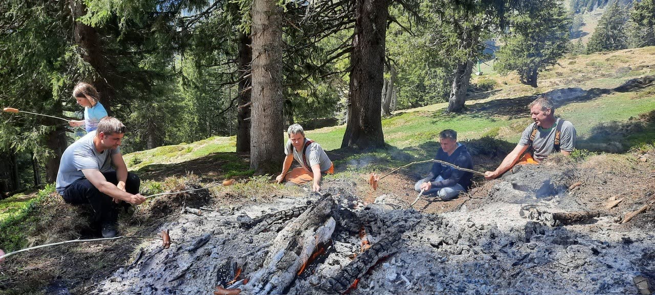 Helfer an Glut-Feuerstelle im lichten Bergwald, Alpweide