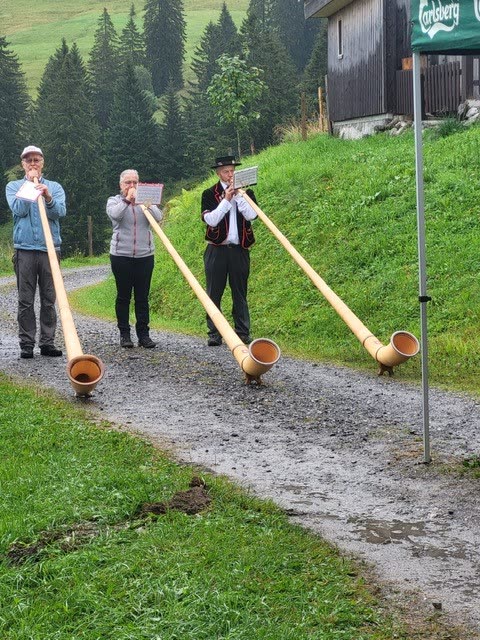 Drei Alphornbläser auf Kiesweg vor Alphütte