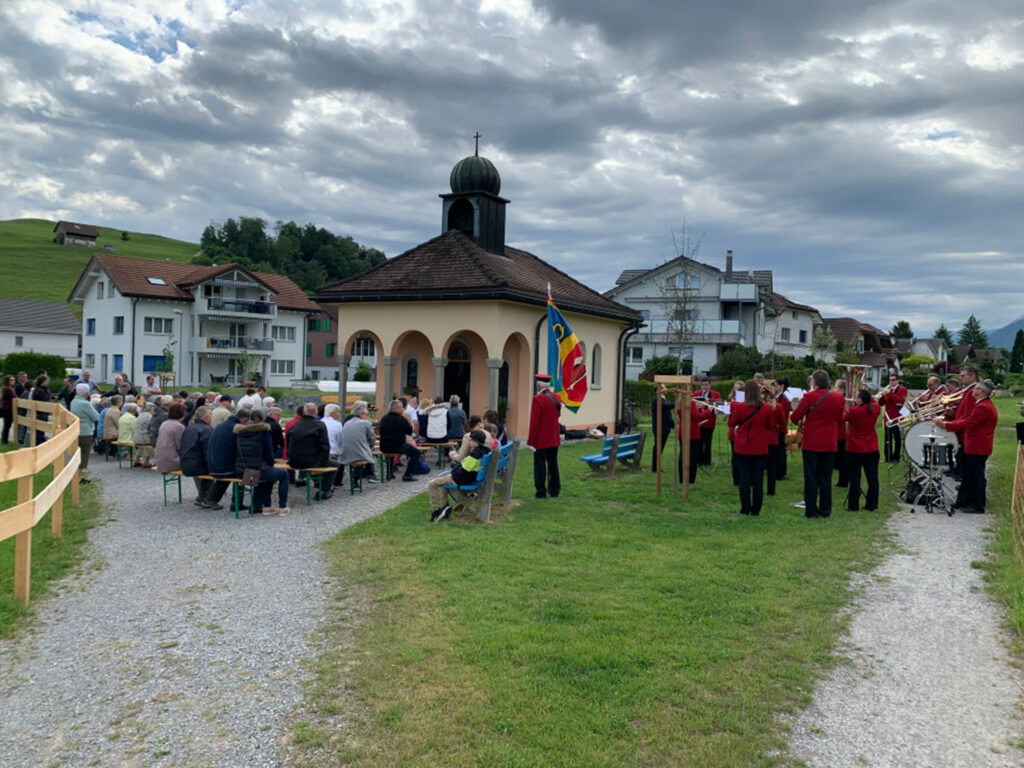 Nuoler Bittgang – St.-Wendelinskapelle Nuolen mit Besuchern, Musikgesellschaft in Rot, bewölkt