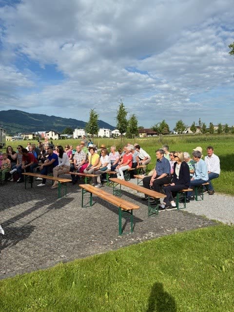 Besucher auf Festbänken auf Platz vor der Kapelle, Wiese und Berge