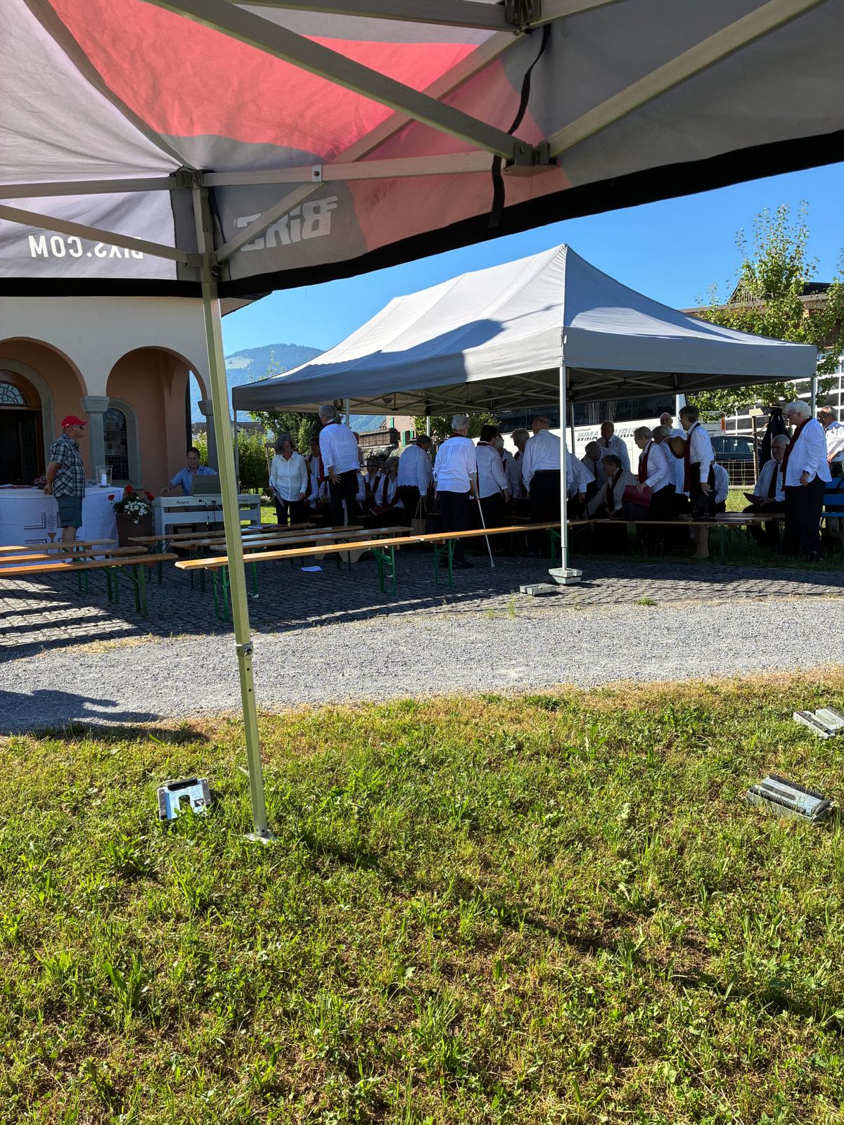 Blick unter Pavillon auf Kapelle und Zelt mit Besuchern, viel Rasen im Vordergrund