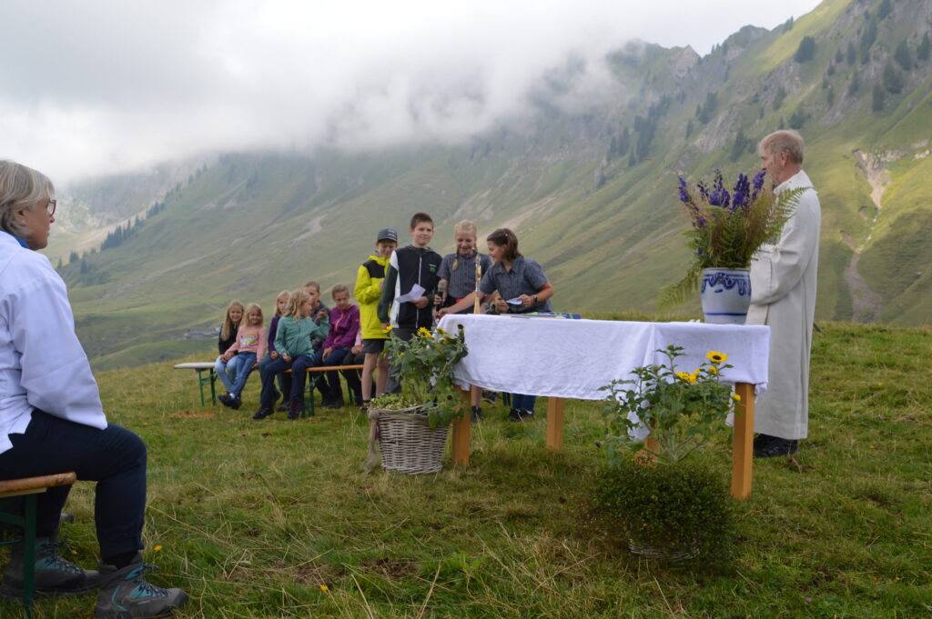 Feldaltar mit Blumen, Pfarrer, Familien auf Bänken, Bergflanke mit Wolken
