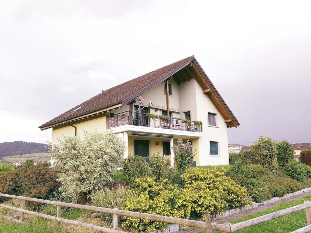 Hellgelbes Wohnhaus mit Satteldach und Balkon an der Falkenstrasse hinter einem blühenden Garten